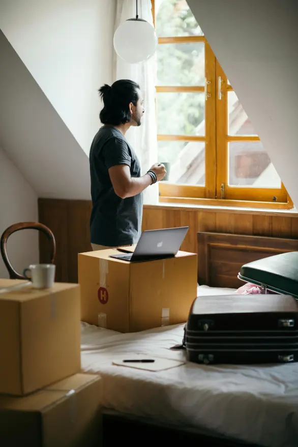 A man looking out a bedroom window as he takes a break from packing boxes